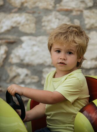 Funny boy car driver with steering wheel. Cute boy in yellow T-shirt in red toy car. Little boy driving big toy car and having fun outdoors. Young kid beautiful portrait active life. Child traveling.の写真素材