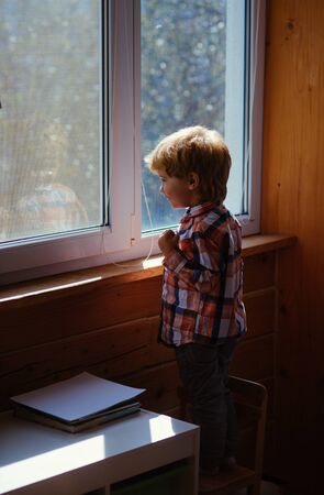 Little boy looks in window with curiosity. Sunlight comes into room through windows. Big white plastic windows full of sunlight. Cute kid enjoy sun at home through windows.の写真素材