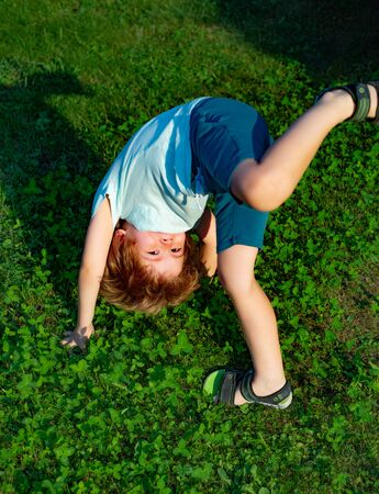 Upside down. The boy is doing exercise on the grass. Equilibrium. The child raises his leg up. Happy childhood with walks and games in nature.の写真素材