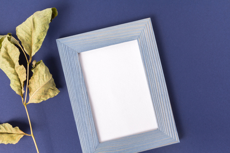 A blue wooden frame and a dried leaf.  paper inside the frame with place for text. Blue-purple background made of pastel embossed paper. Top View. The composition is cropped at an angle.の写真素材