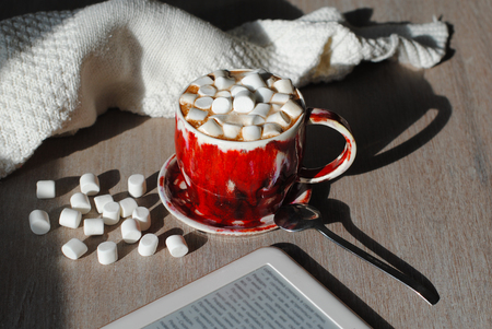 A red cup of cocoa on a gray wooden background with marshmallows and an ebook nearby.の写真素材