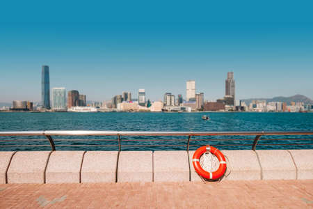 Sidewalk, railing and lifebelt at Victoria Harbor waterfront promenade with Kowloon skyline, HongKongの写真素材