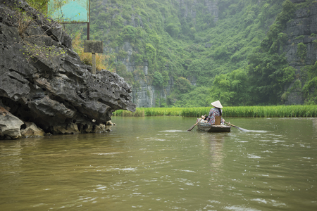 Vietnamese woman in traditional conical hat rows boat into natural cave on Ngo Dong river, Tam Coc, Ninh Binh, Vietnamの写真素材