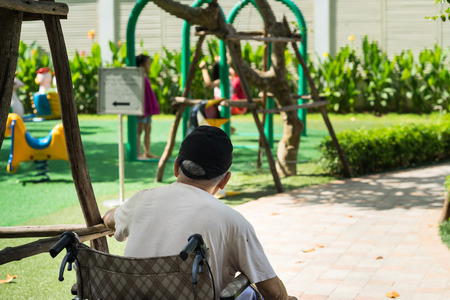 Old man sits on wheelchair looking at children playing on playground. Concept of youth recall, taking care of children.の写真素材