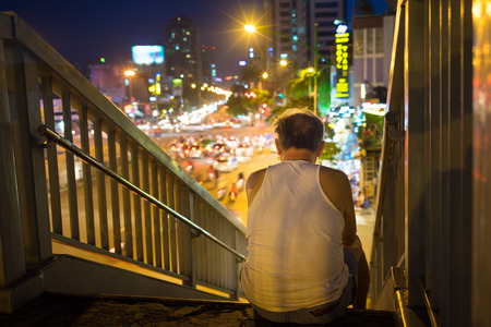 Old man sits on walking bridge seeing crowded city by night. Concept of lonely old manの写真素材