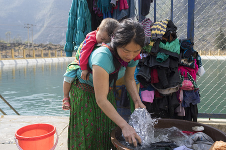Ha Giang, Vietnam - Feb 7, 2014: Unidentified Hmong very young mother washing clothes at a lake, with the son sleeping on her backのeditorial素材