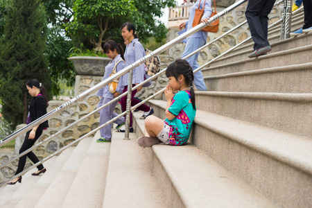 Quang Ninh, Vietnam - Mar 22, 2015: Little girl child clasp her hands praying at Cai Bau old temple in Van Don. Some buddhist on brown clothes on backgroundのeditorial素材