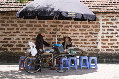 Hanoi, Vietnam - July 16, 2916: Woman who sales stewed fruit, sweetened porridge at stall talking to old man, old brick on background, in Duong Lam ancient village, Son Tay district.のeditorial素材