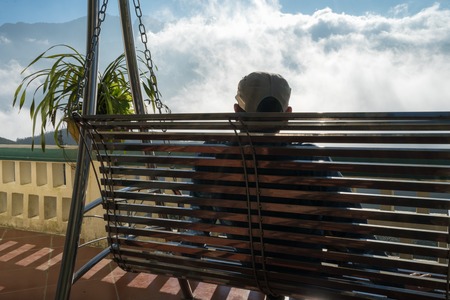 Male tourist sit on swing seeing mountain landscape with low white clouds under brilliant sky. Ideas for travel and feeling.の写真素材