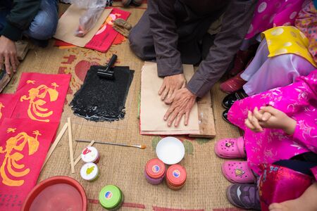 Hanoi, Vietnam - Jun 22, 2017: Old Dong Ho village artist making Dong Ho folk woodcut painting with children surrounded in communal house, Quoc Oai district. Concept of cultural preservationのeditorial素材