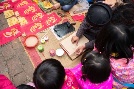 Hanoi, Vietnam - Jun 22, 2017: Old Dong Ho village artist making Dong Ho folk woodcut painting with children surrounded in communal house, Quoc Oai district. Concept of cultural preservationのeditorial素材