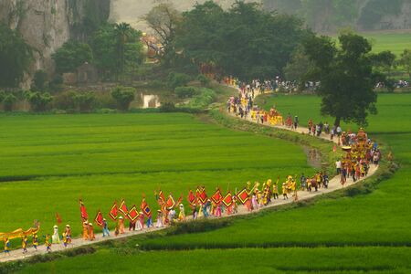 Ninh Binh, Vietnam - Apr 10, 2017: Thai Vi traditional spring festival with crowded people and palanquin, dancing dragon, flag walking on curved soil road among rice fieldのeditorial素材