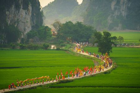Ninh Binh, Vietnam - Apr 10, 2017: Thai Vi traditional spring festival with crowded people and palanquin, dancing dragon, flag walking on curved soil road among rice fieldのeditorial素材