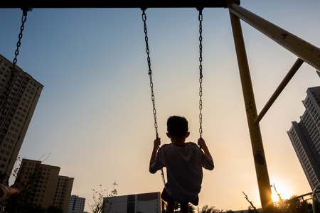 Child swinging on swing in sunset in city with building on backgroundの写真素材