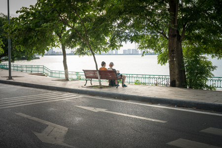 Two young women sitting on bench under big tree by the lake in cityの写真素材