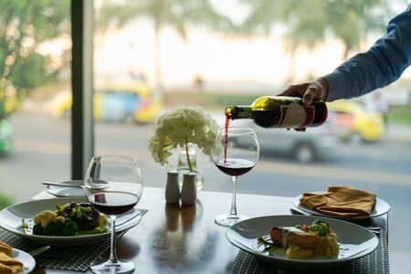 Fresh romantic breakfast table next to morning briliant light window, with beef, salmon fish, mashed potato, vegetable and waiter hand pouring red wineの写真素材