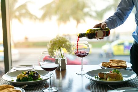 Fresh romantic breakfast table next to morning briliant light window, with beef, salmon fish, mashed potato, vegetable and waiter hand pouring red wine