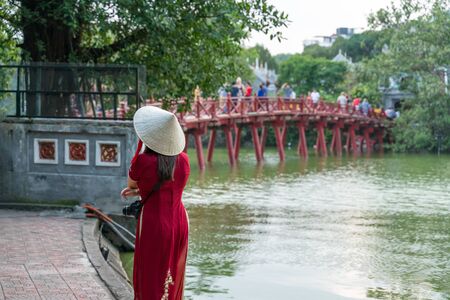 Female tourist wearing Vietnamese traditional dress Ao Dai with The Huc bridge at Hoang Kiem lake on backgroundの写真素材