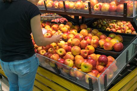 Fresh healthy fruits on shelves in supermarket, with female buyer choosing red applesの写真素材