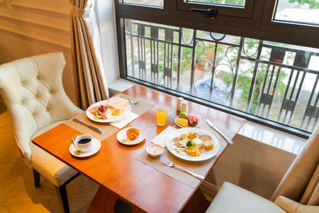 Fresh breakfast table next to window with bread, pastry, egg, fruit, juice, coffee cupの写真素材