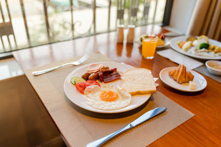 Fresh breakfast table next to window with bread, pastry, egg, fruit, juice, coffee cupの写真素材