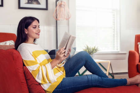 An attractive, young woman enjoys reading a book at home on her red sofa