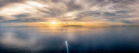 A boat traveling over idyllic, calm ocean towards an lonely island during sunset time with cloudscape in the skyの写真素材