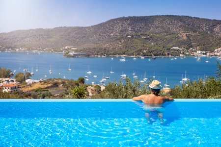 A tourist man in a swimming pool enjoys the view of the sailing boats in a bay at Poros island, Saronic Gulf, Greece, during summer timeの写真素材