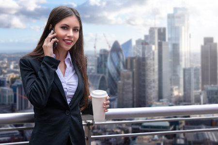 A attractive business woman in a corporate outfit holding a coffee cup talks on her mobile telephone in front of the city skyline of London, Englandの写真素材