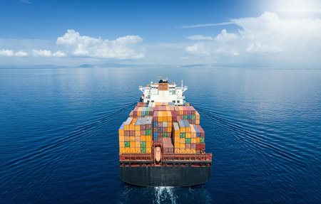 Aerial back view of a large container cargo carrier ship traveling over very calm ocean with sunshineの写真素材