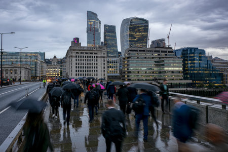 Blurred motion view of commuters with umbrellas in London going to work on a rainy, gray day in front of the city skyline during morning rush hourの写真素材