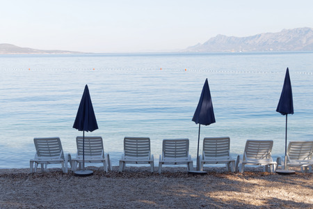Lots of white empty deck chair and blue parasol standing on the beach in the early morning before...