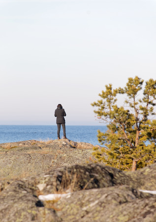Silhouett of a thoughtful woman in black clothes looking at the sea. Rocks and a small pine tree in the foregroundの写真素材