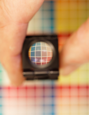 Hand holding a magnifyer or loupe above a colorful test print with color shades in cyan, blue, magenta, red, green, orange, and yellow. Short depth of focusの写真素材