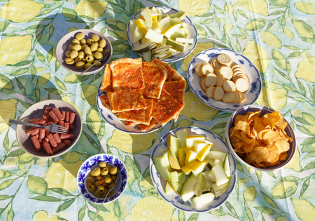 Bowls filled with pizza, melon, olives, potato chips, sausage and crackers on a table with a colorful cloth in yellow and green waiting for the guestsの写真素材