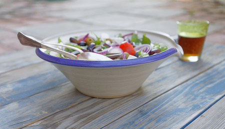 Bowl filled with sallad, tomato, red onion, olives and sheep cheese and a glass of beer on a blue wethered wooden tableの写真素材