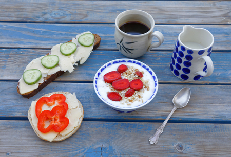 Blue wodden wethered table set for breakfast including coffe, milk, yoghurt with strawberry and cereals, sandwich with cheese, cucumber and red pepperの写真素材