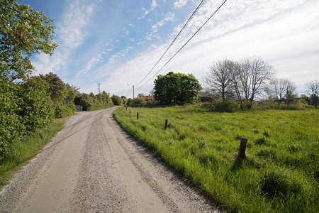 Rural country road in the small village during spring, distant red cottage and trees, blue skyの写真素材