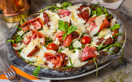 Black pizza with Parma ham, parmesan cheese, dried tomatoes, rocket and basil.の写真素材