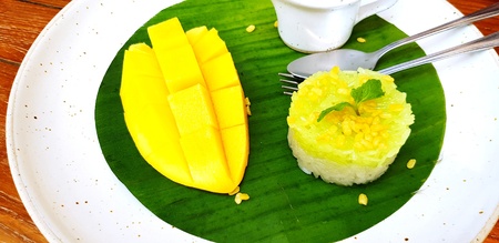 Close up Thai local dessert food with mango, sticky rice, jug of coconut milk, spoon and fork on banana leaf in white plate on wooden table with copy space at cafÃ© - Delicious and Natural materialの写真素材