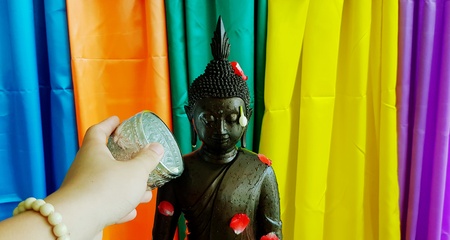 Close up hand splashing or pouring ancient black Buddha statue with petal red rose, Jasmine isolated on colorful curtain background- Songkran festival is Thailand tradition or Culture and respectの写真素材
