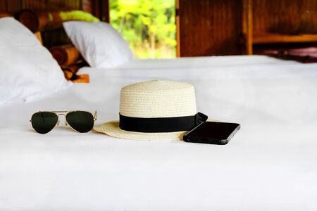 Vacation time on weekend with black sunglasses, white vintage straw hat and smartphone on white bed with copy space. Travel, Accessories, Fashion and Resting time conceptの写真素材