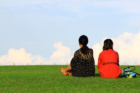 Two Asian women in black and red dress sitting on grass field and relaxing on weekend at garden park with cloud and blue sky background with copy space on left.の写真素材