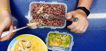 Top view or Flat lay and Close up hand using spoon and fork eating brown rice in plastic dishware with  chicken coconut soup and  Stir fried zucchini with eggs on blue marble table for background.の写真素材