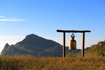 Golden bell hanging on wooden pole on top mountain among glass field for alarm time to people in the morning with big mountain and clear blue sky background at Doi Pha Tang, Chiang Rai, Thailand.の写真素材