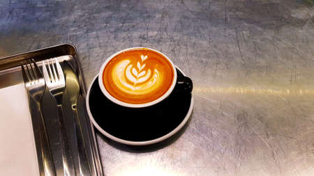 Latte art cappuccino coffee of black cup with white napkin, fork and knife in tray on silver or...