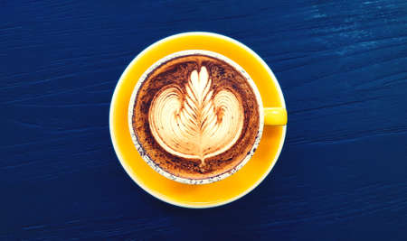 Top view of cup of art cappuccino or latte coffee on dark blue wooden background in vintage tone. Flat lay of hot drinking in yellow glass. Food design concept.の写真素材