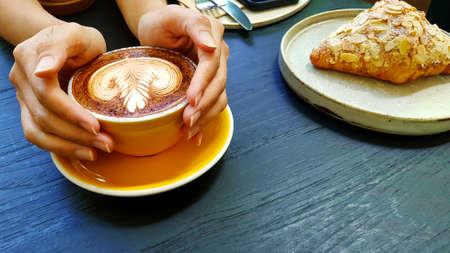 Woman's hand holding yellow cup of art latte coffee with almond cream croissant, knife, fork and...