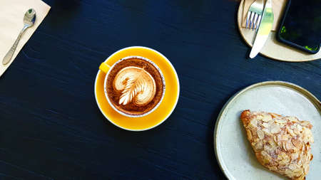 Top view of art latte coffee with almond cream croissant, knife, fork and phone on blue wooden...