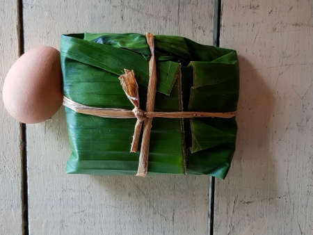Top view of boiled egg and rice wrapped in banana leaves on white wood background. Flat ay of food on hardwood panel. Lunch and use natural material contain instead of plastic, foam box or plate.の写真素材